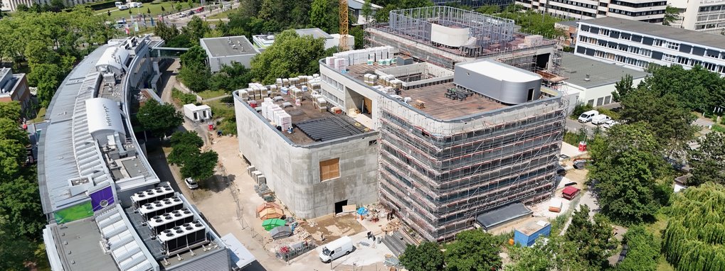 Luftbild eines im Bau befindlichen Gebäudekomplexes, bestehend aus mehreren miteinander verbundenen Strukturen, umgeben von Bäumen und anderen Gebäuden.