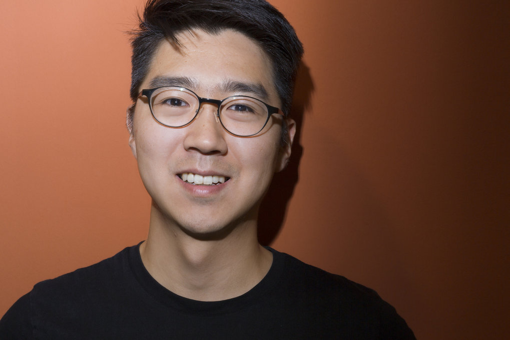 An Asian-looking young man with glasses and a black T-shirt stands in front of an orange background and smiles into the camera.